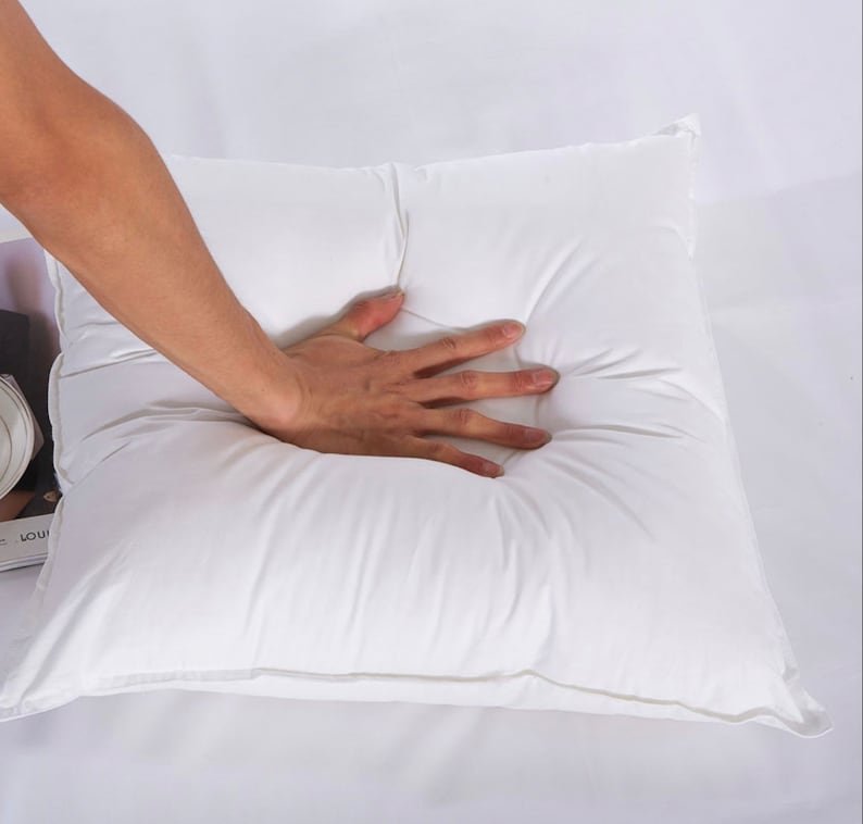 May include: A white square pillow with a hand pressing down on the centre of the pillow.