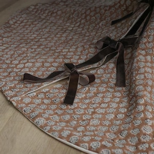 May include: A round, brown tree skirt with a repeating floral pattern in white. The skirt has a white border and is adorned with dark brown velvet ribbon ties.