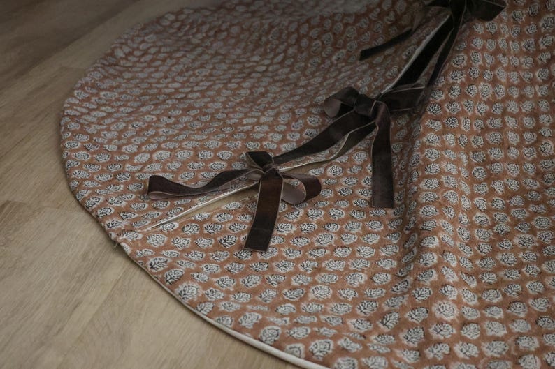 May include: A round, brown tree skirt with a repeating floral pattern in white. The skirt has a white border and is adorned with dark brown velvet ribbon ties.