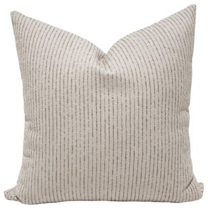 May include: A square throw pillow featuring a textured, light beige fabric base with a repeating pattern of thin, vertical dark gray stripes. The pillow has a soft, plush appearance with a visible crease at the top center, creating a structured shape.