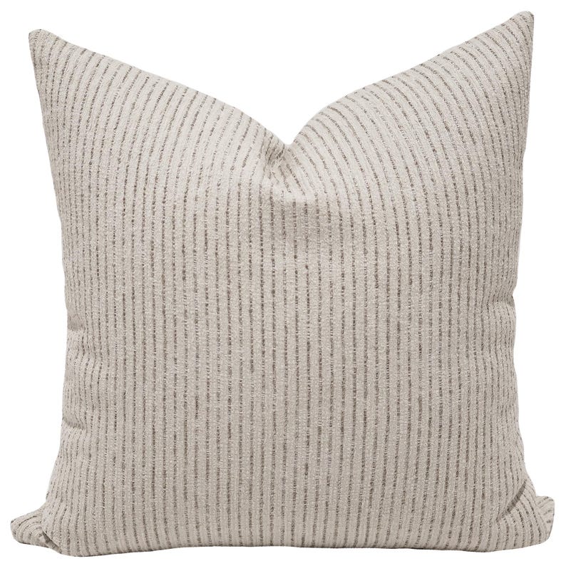 May include: A square throw pillow featuring a textured, light beige fabric base with a repeating pattern of thin, vertical dark gray stripes. The pillow has a soft, plush appearance with a visible crease at the top center, creating a structured shape.