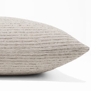 May include: A light beige throw pillow featuring a textured, horizontal ribbed pattern across its surface. The fabric displays a subtle variation in tone with darker flecks integrated into the weave, creating a consistent, linear appearance.