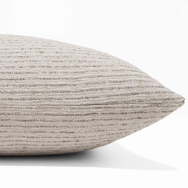 May include: A light beige throw pillow featuring a textured, horizontal ribbed pattern across its surface. The fabric displays a subtle variation in tone with darker flecks integrated into the weave, creating a consistent, linear appearance.