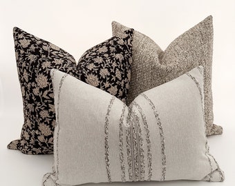 May include: Three decorative throw pillows with different patterns. The pillow in the front is a light beige with a textured pattern of vertical lines. The pillow in the back is a black and cream floral print. The pillow in the middle is a light beige with a textured pattern.