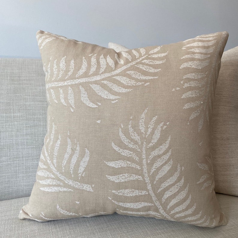 May include: Beige square throw pillow with a white fern leaf pattern. The pillow is made of a textured fabric and is placed on a light gray sofa. The fern leaf design is repeated across the pillow's surface, creating a natural, botanical theme.