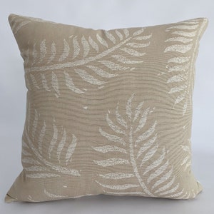 May include: Square beige throw pillow with a repeating pattern of off-white fern leaves. The pillow has a textured appearance and is likely made of fabric. The design is a neutral color palette, suitable for various home decor styles.