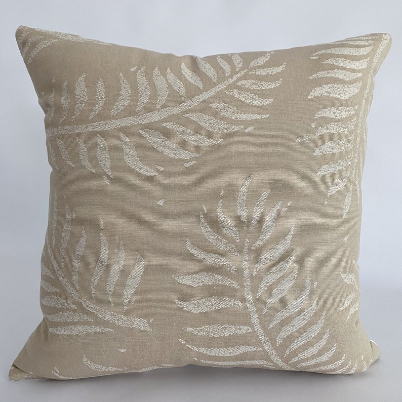 May include: Square beige throw pillow with a repeating pattern of off-white fern leaves. The pillow has a textured appearance and is likely made of fabric. The design is a neutral color palette, suitable for various home decor styles.