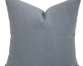 Op de afbeelding: Een vierkant kussen in een gedempte blauwgrijze tint. Het kussen heeft een zachte, getextureerde uitstraling, wat wijst op linnen of een vergelijkbare stof. Het is een decoratief item voor een bed of bank.