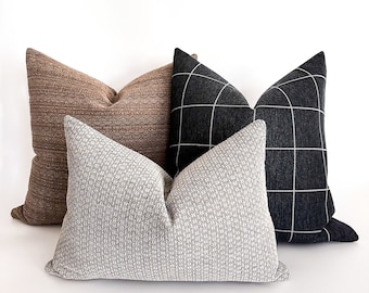 May include: Three decorative throw pillows in a variety of textures and patterns. The pillows are arranged in a stack, with the largest pillow on the bottom and the smallest pillow on top. The bottom pillow is a brown and beige striped pillow. The middle pillow is a black and white checked pillow. The top pillow is a white and grey textured pillow.