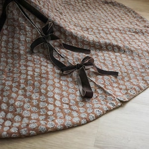 May include: A brown and white floral patterned tree skirt with dark brown velvet ribbon ties. The skirt is round with a white border and is made of a soft fabric. The design features a repeating pattern of small white flowers.
