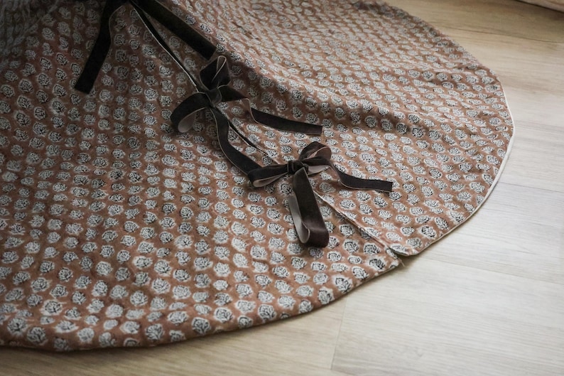 May include: A brown and white floral patterned tree skirt with dark brown velvet ribbon ties. The skirt is round with a white border and is made of a soft fabric. The design features a repeating pattern of small white flowers.