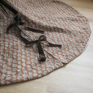 May include: A round, brown tree skirt with a white floral pattern and a white border. The skirt has a split opening and is tied with dark brown velvet ribbons. The fabric has a soft, textured appearance.