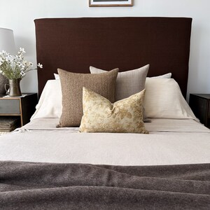 Op de afbeelding: Een bed met een bruin hoofdbord en een wit linnen dekbedovertrek. Er liggen vier kussens op het bed, twee bruin, een wit en een met een bloemenpatroon. Het bed is gemaakt met een bruin wollen deken.