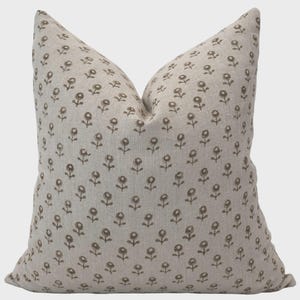 May include: A beige throw pillow with a repeating pattern of small, brown floral designs. The pillow has a soft, textured appearance and is likely made of linen or a similar fabric. The floral pattern adds a touch of rustic charm.