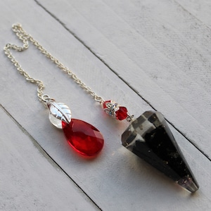 May include: A silver chain dangles two pendulums. One is a teardrop-shaped red crystal with a silver leaf charm. The other is a black and clear crystal point with a silver bead and red crystal accents.