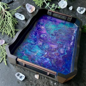 May include: A rectangular black wooden tray with a swirling resin design in blue, teal, and purple. The tray has bronze handles. Crystals and greenery surround the tray. The tray is a decorative item.