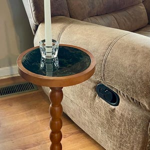 May include: A small, wooden side table with a round top featuring a dark green marble-like design. A clear glass candle holder with a white candle sits on top. The table has a turned, bulbous stem and a round base.