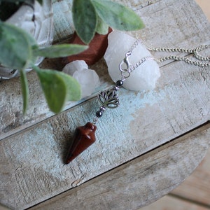 May include: A brown crystal pendulum with a silver chain and a silver lotus charm. The pendulum is hanging from a white surface.