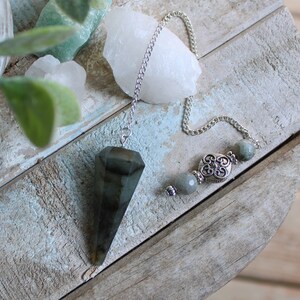 May include: A silver chain with a dark gray crystal pendulum and gray beads. The pendulum is a pointed, triangular shape.