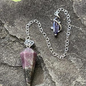 May include: A pendulum featuring a faceted, conical-shaped stone with a gradient of deep magenta and dark gray. The stone is attached to a silver-toned metal chain with a Celtic knot connector and a small, purple, marquise-cut crystal charm.