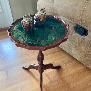 May include: A small, round, wooden side table with a dark finish. The tabletop features a green and gold marbled design and is adorned with two owl figurines. The table has a three-legged base.