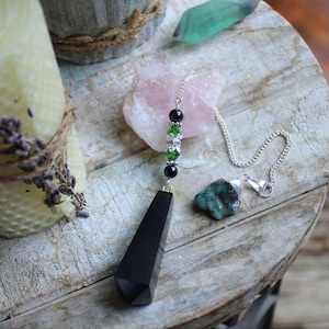 May include: A black obsidian pendulum with a silver chain and decorative beads. The beads include black and green crystals. An emerald and other crystals are also present. The pendulum is displayed on a wooden surface.