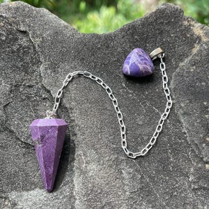 May include: A purple faceted stone pendulum with a pointed base attached to a silver-colored metal chain. The chain connects to a small, triangular purple stone piece at the opposite end, featuring visible white veining throughout the stone surfaces.