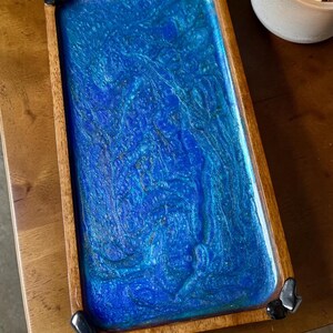 May include: A rectangular wooden tray featuring a vibrant, swirling blue and teal resin inlay with a glossy finish. The tray is accented with small, smooth black stones attached to each of the four corners, providing a distinct decorative edge.