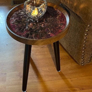 May include: A round wooden side table with a dark stained finish and black legs. The tabletop features a resin design with shades of purple, brown, and white. A clear glass candle holder with a lit candle sits on top.