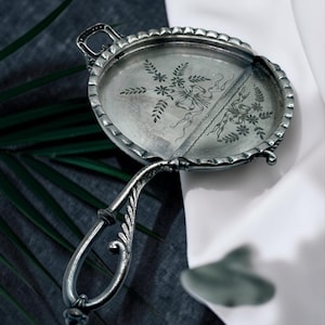 May include: A silver-toned metal hand mirror with an oval frame featuring a scalloped edge. The reflective surface is divided by a vertical line and decorated with an etched floral motif of leaves and ribbons. It has a long, ornate handle.