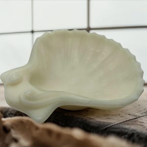 May include: A creamy off-white glass dish shaped like a scallop shell. The bowl features a smooth, translucent finish with subtle ridges radiating from the centre toward the scalloped edge, and a decorative curled detail at the base hinge.