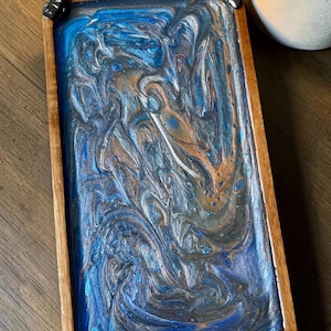 Può includere: Un vassoio rettangolare in legno con un inserto in resina colata caratterizzato da un motivo marmorizzato di blu profondo, rame metallizzato e vortici argentati. Il vassoio è decorato con quattro piccole pietre nere lisce su ogni angolo.