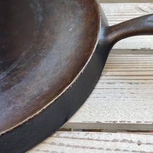May include: Close-up of a well-used, dark brown cast iron frying pan with a long handle. The pan's interior shows signs of use, with a smooth, slightly textured surface. The exterior is black, and the handle is integrated into the pan's design.