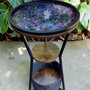 May include: A round side table with a black metal frame and a decorative top. The tabletop features a swirling pattern of purple, blue, and white. The table has a wooden shelf and a gray fabric basket. The table is approximately 61 cm tall.