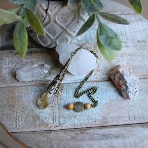 May include: A clear quartz crystal pendulum with a brass chain and beads. The pendulum is hanging over a wooden surface with a white crystal and a pink crystal.