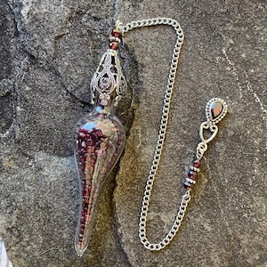 May include: A clear glass teardrop-shaped pendant filled with small dark red garnet beads. The pendant features an ornate silver-toned metal cap and is attached to a silver-toned chain with additional red bead accents and a teardrop charm.