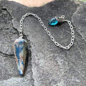 May include: A faceted, cone-shaped pendulum made of blue and brown stone, attached to a silver-colored metal chain. The chain features a small, translucent blue teardrop-shaped bead at the end, secured with a decorative silver-colored metal cap.