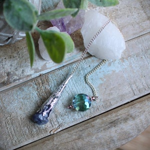 May include: A silver pendulum with a green crystal point and a silver chain. The pendulum is resting on a wooden surface.