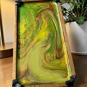 May include: A rectangular wooden tray with a vibrant, swirling epoxy resin design in shades of green, yellow, and brown. The tray has a natural wood frame and black corner accents. The tray is a decorative item.