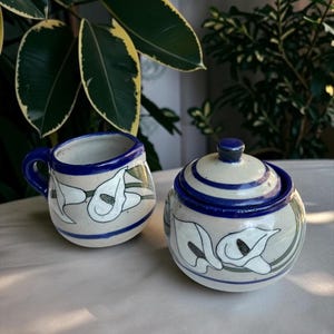 May include: A ceramic mug and a matching lidded sugar bowl, both featuring a cream-colored base with hand-painted white calla lily flowers, green stems, and bold cobalt blue rim accents. The pieces have a rounded shape and a glossy finish.