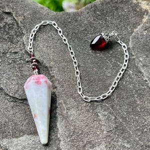 May include: A pendulum featuring a faceted, conical white stone with pink inclusions, attached to a silver-toned metal chain. The chain includes small red beads and terminates in a dark red, teardrop-shaped stone charm with a decorative silver cap.