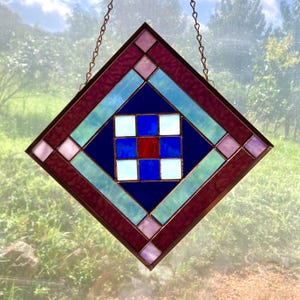 May include: A diamond-shaped stained glass suncatcher featuring a central nine-patch grid of white, blue, and red glass squares. The center is surrounded by dark blue, light blue, and deep red textured glass borders with light pink corner accents.