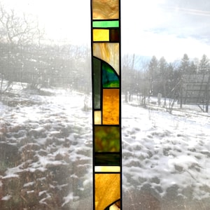May include: A rectangular stained glass panel with a geometric design. The panel features a mix of colors, including gold, green, yellow, and black. The glass is divided into various shapes and sizes, creating an abstract pattern. It is suspended by a chain.