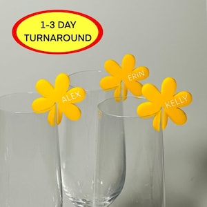 May include: Three yellow flower-shaped glass markers with the names Alex, Erin, and Kelly written in white. The markers are placed on three clear glass champagne flutes.