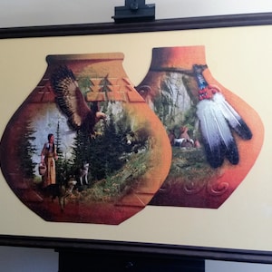 Puede incluir: Una obra de arte enmarcada que presenta dos vasijas de cerámica estilizadas. La vasija de la izquierda representa a una mujer en un bosque con un pájaro volando por encima. La vasija de la derecha presenta una pluma blanca con puntas negras.