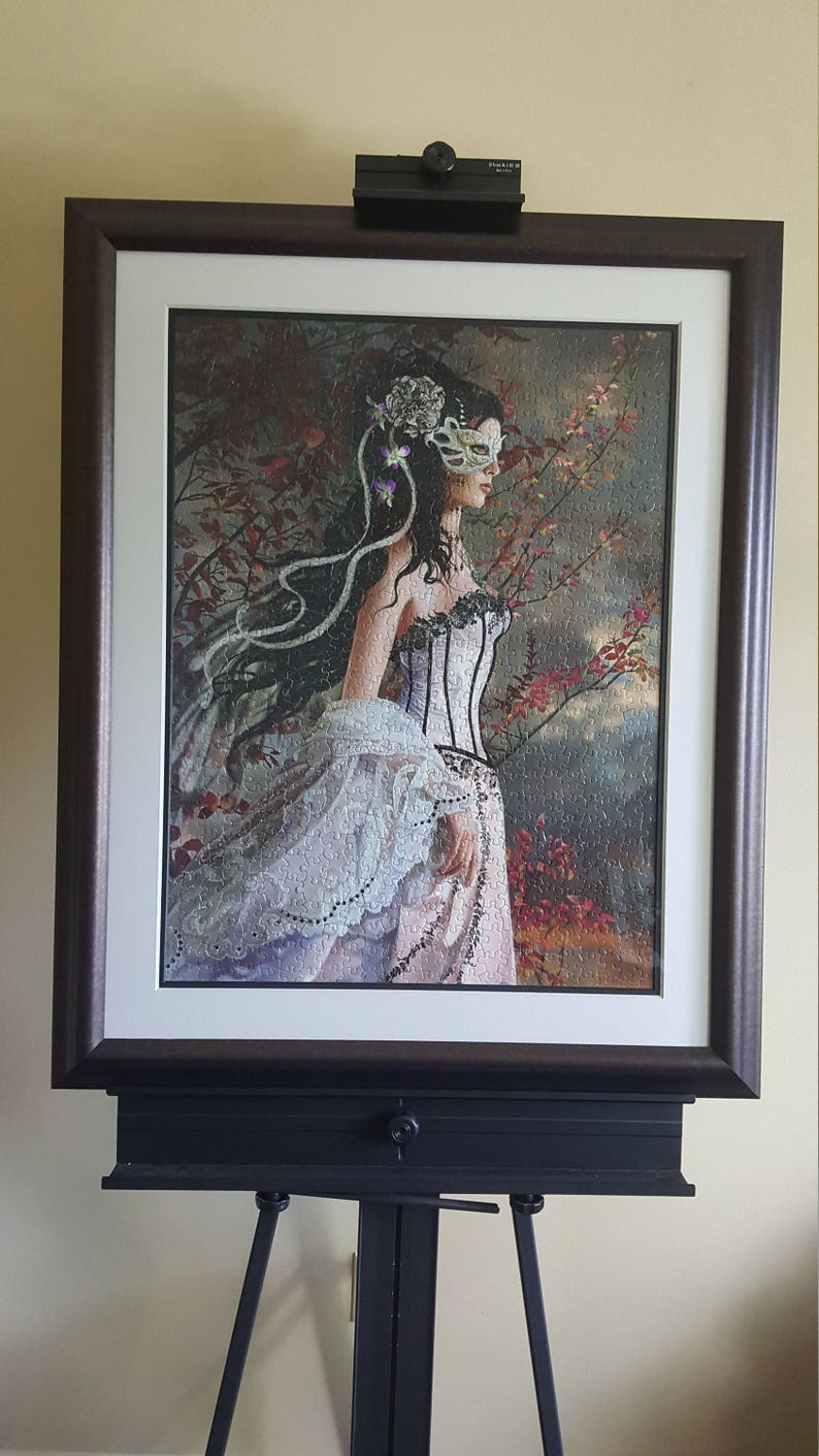 K&ouml;nnte beinhalten: Ein gerahmtes Puzzle, das eine Frau in einem wei&szlig;en Kleid und einer Maskenballmaske zeigt, die in einer Waldlandschaft mit Herbstlaub steht.