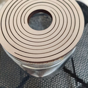 May include: A wooden lid with a circular design featuring concentric rings. The lid has a hole in the center and is made of light brown wood.