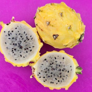 May include: Three yellow dragon fruit halves with black seeds on a pink surface. The fruit is cut in half and the inside is white with black seeds.