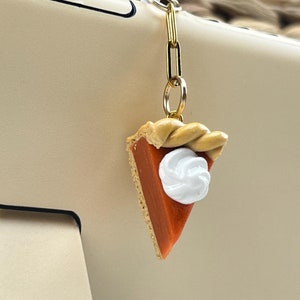 May include: A gold chain with a small, handmade pumpkin pie charm. The pie is orange with a white swirl of whipped cream on top. The charm is attached to a clear phone case.