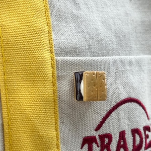 May include: A beige canvas tote bag with a vertical yellow stripe and a s'more-shaped pin attached to the front pocket. The bag features the red embroidered text 'TRADE JOE'S' partially visible at the bottom right corner of the front pocket.
